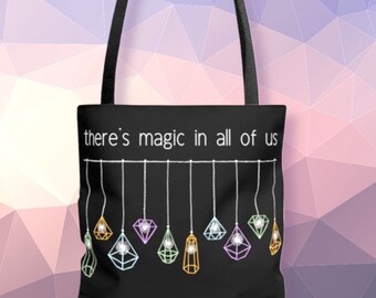 May include: Black tote bag with the text "there's magic in all of us" in white. The bag features a design of hanging geometric shapes in various colors, including green, pink, and yellow. The bag has black handles.