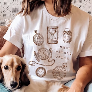 Puede incluir: Camiseta blanca con un gr&aacute;fico marr&oacute;n de varios relojes y el texto "amor vincit omnia".