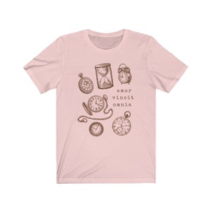 Puede incluir: Una camiseta rosa con un gr&aacute;fico de varios relojes y un reloj de arena. El texto "amor vincit omnia" est&aacute; impreso debajo de los relojes.