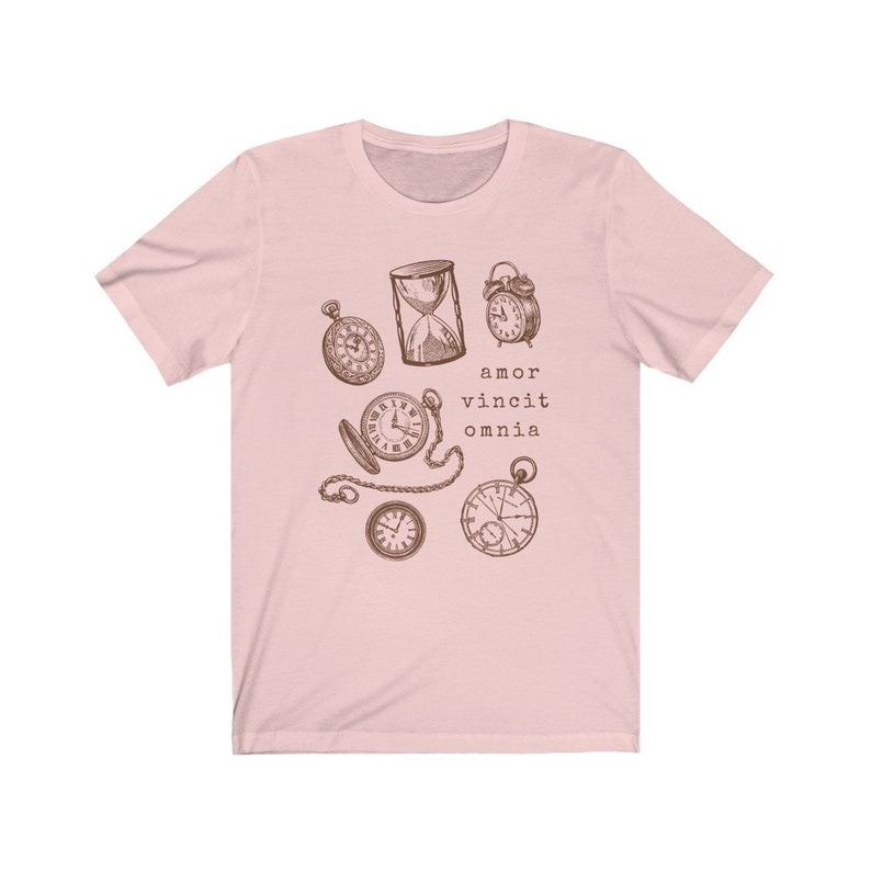 Puede incluir: Una camiseta rosa con un gr&aacute;fico de varios relojes y un reloj de arena. El texto "amor vincit omnia" est&aacute; impreso debajo de los relojes.
