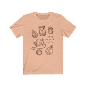 Puede incluir: Una camiseta de color melocot&oacute;n con un gr&aacute;fico de varios relojes y un reloj de arena. El texto "amor vincit omnia" est&aacute; impreso debajo del gr&aacute;fico.