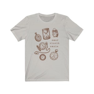 Puede incluir: Una camiseta gris claro con un gr&aacute;fico marr&oacute;n de varios relojes y un reloj de arena. El texto "amor vincit omnia" est&aacute; impreso debajo del gr&aacute;fico.