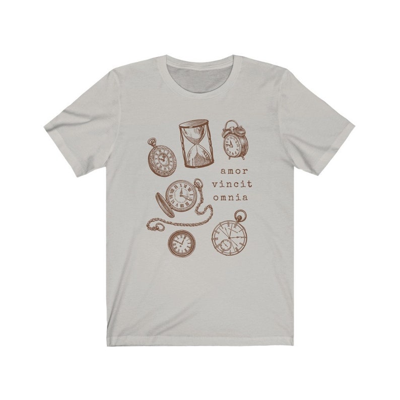 Puede incluir: Una camiseta gris claro con un gr&aacute;fico marr&oacute;n de varios relojes y un reloj de arena. El texto "amor vincit omnia" est&aacute; impreso debajo del gr&aacute;fico.