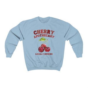 May include: Light blue sweatshirt with the text "CHERRY APOTHECARY" in red and white lettering. A watercolor illustration of two red cherries with green leaves is below the text. The text "LOCALLY SOURCED" is below the cherries.