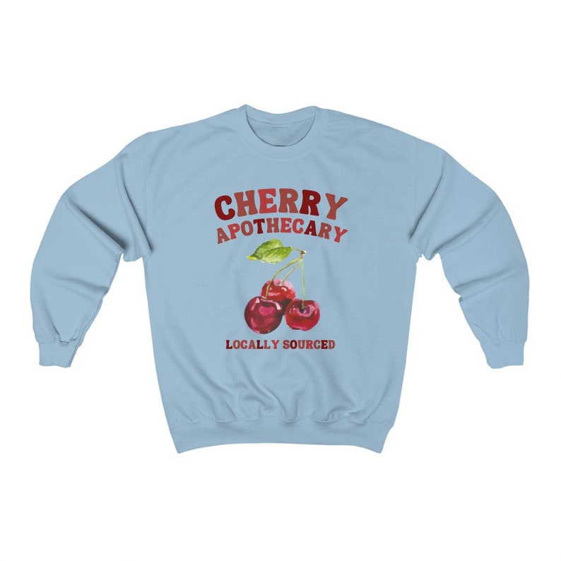 May include: Light blue sweatshirt with the text "CHERRY APOTHECARY" in red and white lettering. A watercolor illustration of two red cherries with green leaves is below the text. The text "LOCALLY SOURCED" is below the cherries.