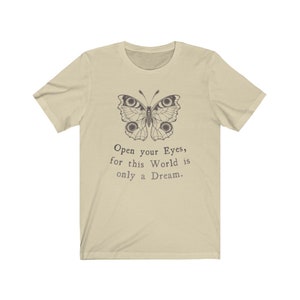 May include: A light beige t-shirt featuring a detailed butterfly illustration in dark brown. Below the butterfly, the text reads, "Open your Eyes, for this World is only a Dream." The shirt has short sleeves and a crew neck.