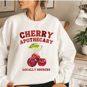 May include: White sweatshirt with the text "CHERRY APOTHECARY" and a graphic of three red cherries with green leaves. The text "LOCALLY SOURCED" is below the cherries.