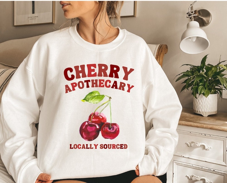 May include: White sweatshirt with the text "CHERRY APOTHECARY" and a graphic of three red cherries with green leaves. The text "LOCALLY SOURCED" is below the cherries.