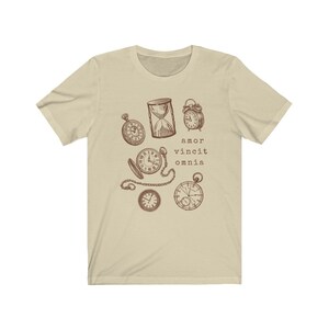 Puede incluir: Una camiseta beige con un gr&aacute;fico marr&oacute;n de varios relojes y un reloj de arena. El texto "amor vincit omnia" est&aacute; impreso debajo del gr&aacute;fico.