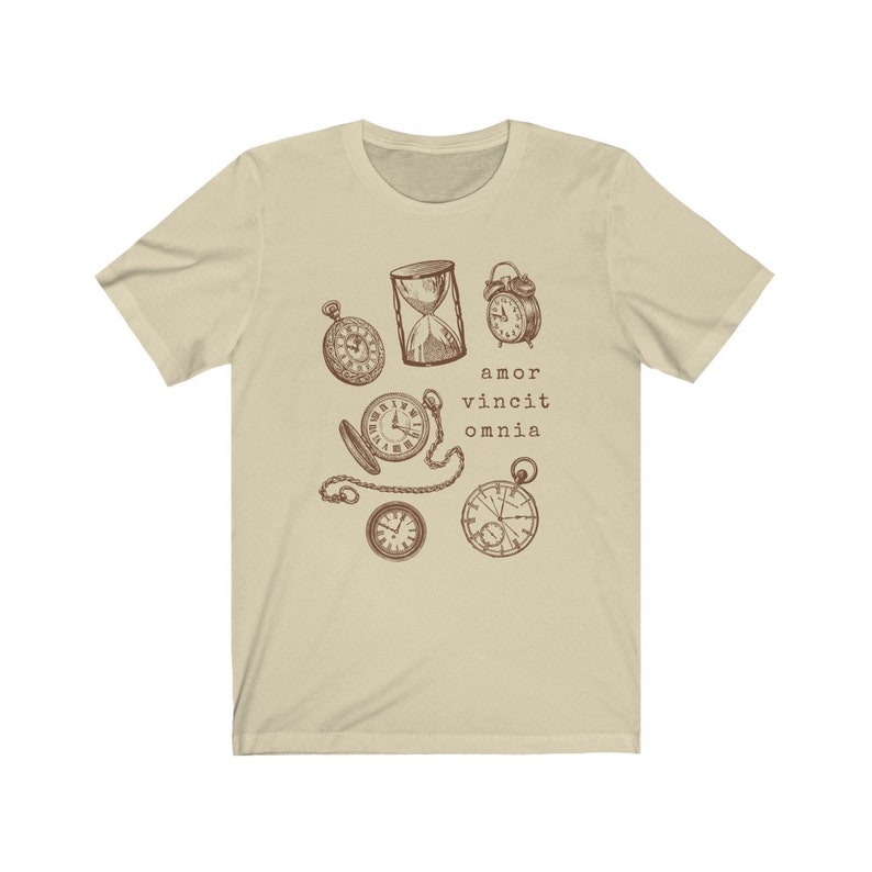 Puede incluir: Una camiseta beige con un gr&aacute;fico marr&oacute;n de varios relojes y un reloj de arena. El texto "amor vincit omnia" est&aacute; impreso debajo del gr&aacute;fico.