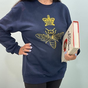 Puede incluir: Una sudadera azul marino con un diseño de corona y abeja dorada. La sudadera está siendo sostenida por una persona que también sostiene un libro titulado "Atlas Obscura".