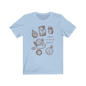 Puede incluir: Camiseta azul claro con un gr&aacute;fico marr&oacute;n de varios relojes y un reloj de arena. El texto "amor vincit omnia" est&aacute; impreso debajo del gr&aacute;fico.
