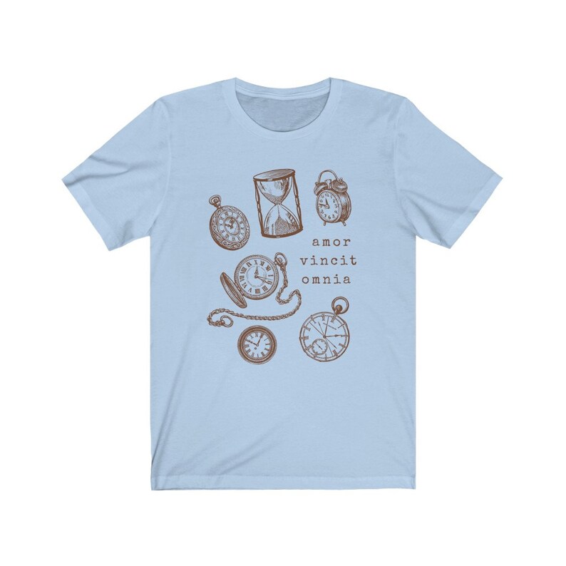 Puede incluir: Camiseta azul claro con un gr&aacute;fico marr&oacute;n de varios relojes y un reloj de arena. El texto "amor vincit omnia" est&aacute; impreso debajo del gr&aacute;fico.
