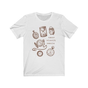 Puede incluir: Camiseta blanca con una ilustraci&oacute;n marr&oacute;n de varios relojes y un reloj de arena. El texto "amor vincit omnia" est&aacute; impreso debajo de la ilustraci&oacute;n.