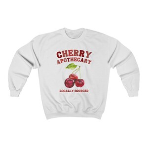 May include: White sweatshirt with a red and white graphic of two cherries with green leaves and the text "CHERRY APOTHECARY LOCALLY SOURCED".