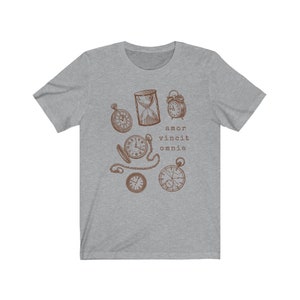 Puede incluir: Una camiseta gris jaspeada con un gr&aacute;fico marr&oacute;n de varios relojes, incluyendo un reloj de arena, relojes de bolsillo y un despertador. El texto "amor vincit omnia" est&aacute; impreso debajo del gr&aacute;fico.