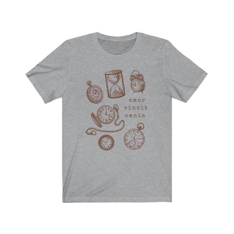 Puede incluir: Una camiseta gris jaspeada con un gr&aacute;fico marr&oacute;n de varios relojes, incluyendo un reloj de arena, relojes de bolsillo y un despertador. El texto "amor vincit omnia" est&aacute; impreso debajo del gr&aacute;fico.