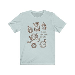 Puede incluir: Camiseta azul claro con un gr&aacute;fico marr&oacute;n de varios relojes y un reloj de arena. El texto "amor vincit omnia" est&aacute; impreso debajo del gr&aacute;fico.