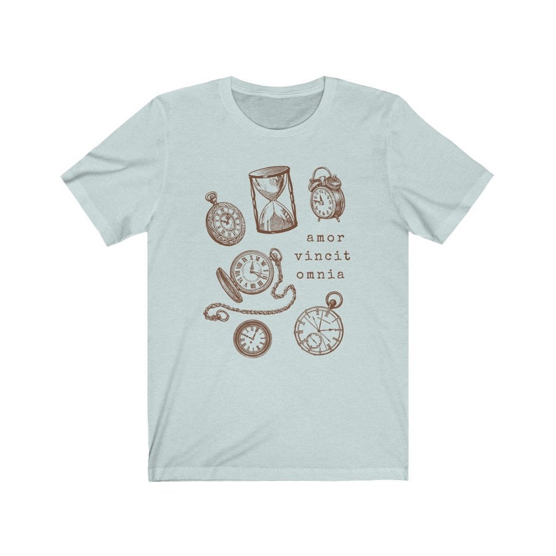 Puede incluir: Camiseta azul claro con un gr&aacute;fico marr&oacute;n de varios relojes y un reloj de arena. El texto "amor vincit omnia" est&aacute; impreso debajo del gr&aacute;fico.