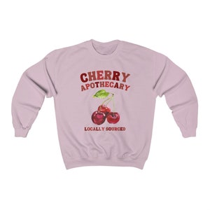 May include: Pink sweatshirt with the text "CHERRY APOTHECARY" in red and orange lettering.  Below the text is a watercolor illustration of two red cherries with green leaves and stems. The text "LOCALLY SOURCED" is below the cherries.