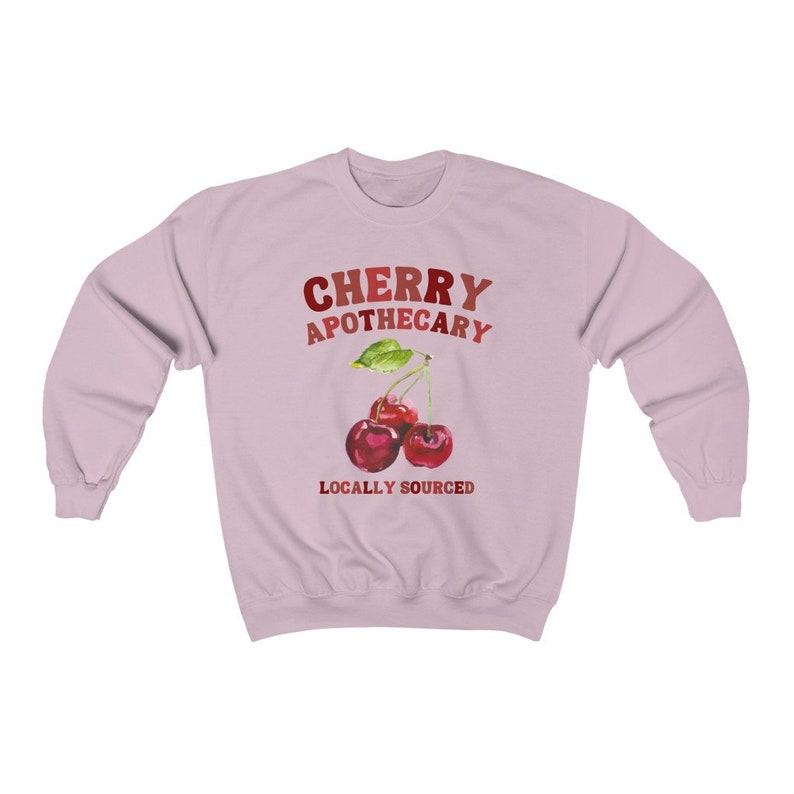 May include: Pink sweatshirt with the text "CHERRY APOTHECARY" in red and orange lettering.  Below the text is a watercolor illustration of two red cherries with green leaves and stems. The text "LOCALLY SOURCED" is below the cherries.
