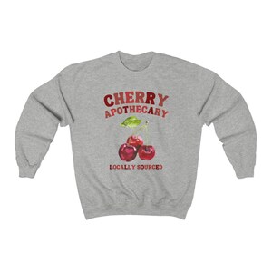 May include: Gray sweatshirt with the text "CHERRY APOTHECARY LOCALLY SOURCED" in red and white. A watercolor illustration of three red cherries with green leaves is centered on the sweatshirt.