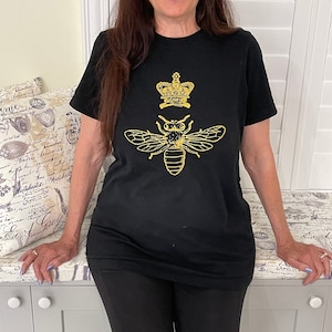 Puede incluir: Camiseta negra con un diseño de corona y abeja dorados.