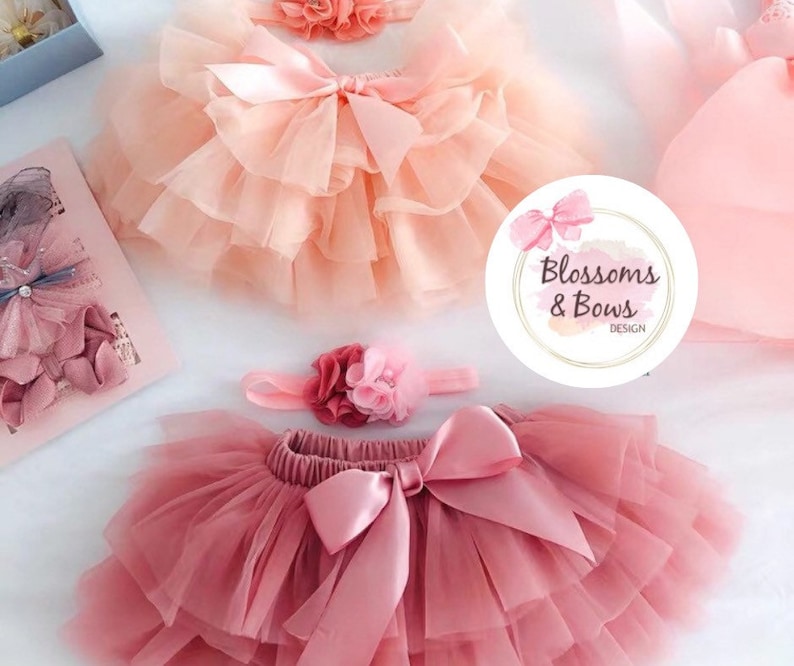 May include: Two tulle tutus, one peach and one dusty rose, with matching satin bows and flower headbands. The tutus are layered and have a soft, delicate look. The headbands are adorned with small, pink flowers. The image also includes a logo for "Blossoms & Bows Design".
