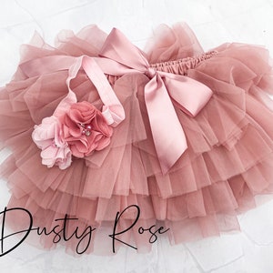 May include: Dusty rose tulle tutu skirt with a matching headband. The skirt has multiple layers of tulle and a satin bow. The headband has a pink flower with a pearl embellishment.