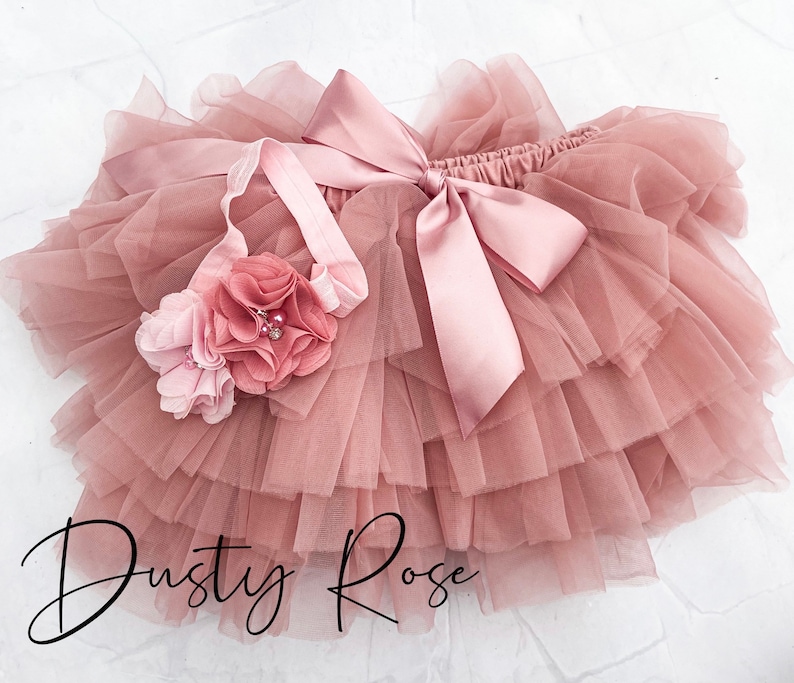 May include: Dusty rose tulle tutu skirt with a matching headband. The skirt has multiple layers of tulle and a satin bow. The headband has a pink flower with a pearl embellishment.