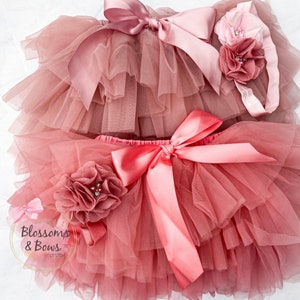 May include: A dusty rose tulle tutu with a pink satin bow and a matching dusty rose flower with a rhinestone center. The tutu has multiple layers of tulle and is perfect for a baby girl.