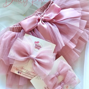 May include: Dusty rose tulle tutu skirt with a satin bow and two matching dusty rose tulle hair bows. The hair bows are packaged on a white card with the text "Blossoms & Bows".