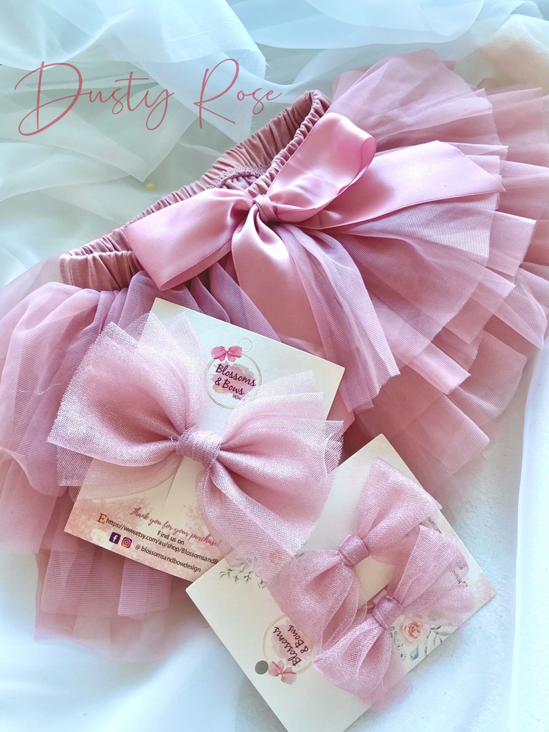 May include: Dusty rose tulle tutu skirt with a satin bow and two matching dusty rose tulle hair bows. The hair bows are packaged on a white card with the text "Blossoms & Bows".