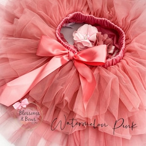 May include: A pink tulle tutu with a large pink satin bow and a pink flower embellishment. The tutu is labeled "Watermelon Pink" and "Blossoms & Bows Design".