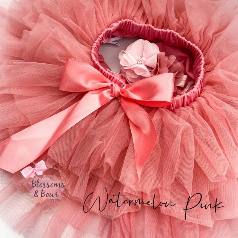 May include: A pink tulle tutu with a large pink satin bow and a pink flower embellishment. The tutu is labeled "Watermelon Pink" and "Blossoms & Bows Design".