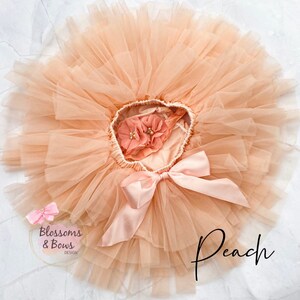 May include: Peach tulle tutu with a pink satin bow and a peach flower embellishment. The tutu is layered and full, with a soft, delicate texture. The text "Peach" is written in black on the bottom right of the image.