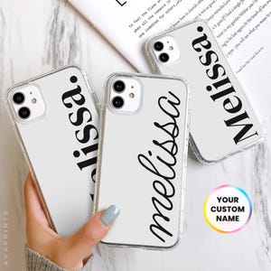 May include: Three clear phone cases with white backgrounds and black text. The text on the cases reads "Melissa" in different styles. The cases are on a white marble surface.