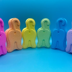 May include: Six bath bombs shaped like cat butts in a rainbow of colors: pink, orange, yellow, green, blue, and purple.