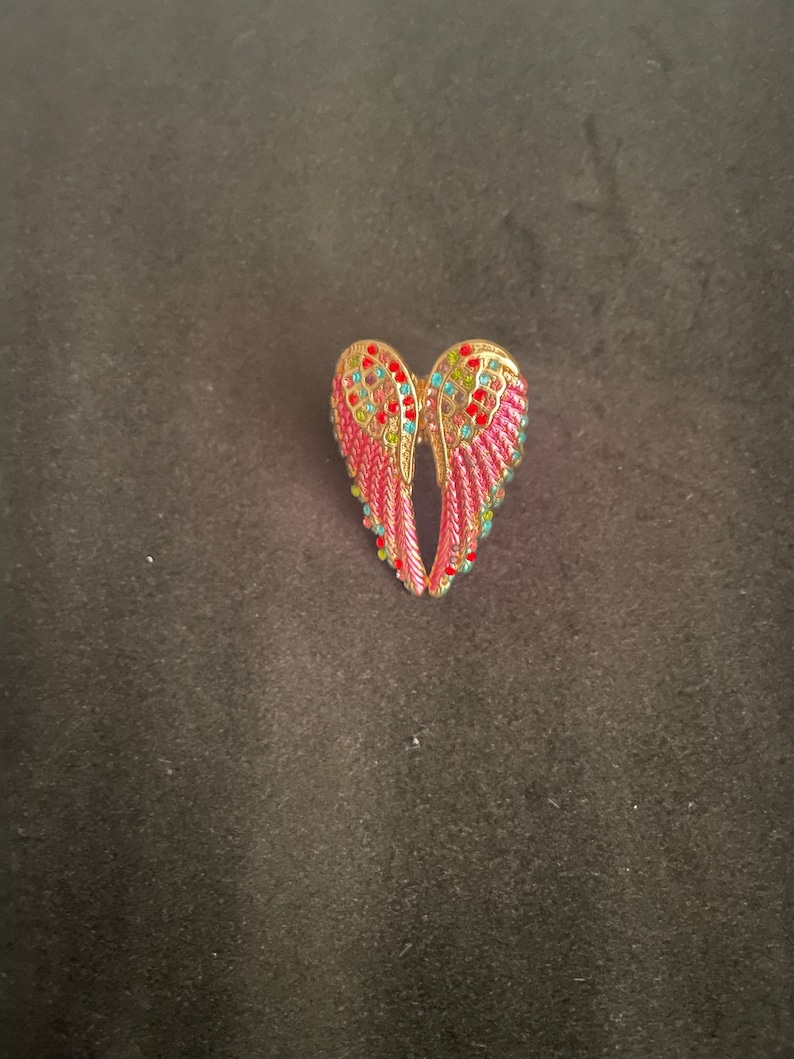 K&ouml;nnte beinhalten: Ein Paar rosa Engelsfl&uuml;gel mit goldenen Verzierungen und bunten Strasssteinen.