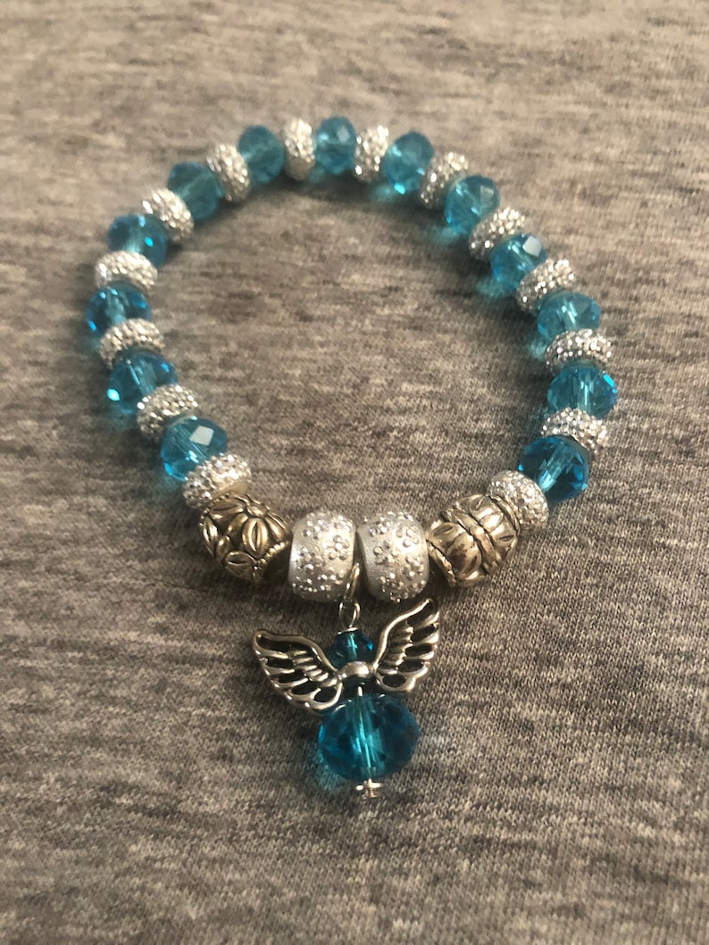 K&ouml;nnte beinhalten: Ein blaues und silbernes Perlenarmband mit einem silbernen Engelsanh&auml;nger. Die Perlen sind facettiert und haben eine glitzernde Oberfl&auml;che.