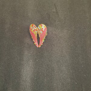 K&ouml;nnte beinhalten: Eine goldfarbene Engelsfl&uuml;gel-Brosche mit rosa und mehrfarbigen Strasssteinen.