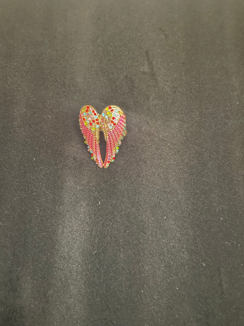 K&ouml;nnte beinhalten: Eine goldfarbene Engelsfl&uuml;gel-Brosche mit rosa und mehrfarbigen Strasssteinen.