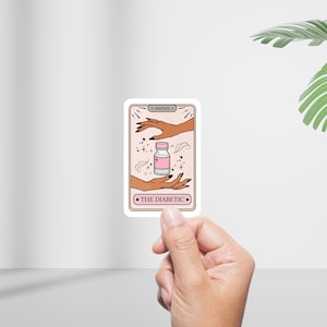 May include: A sticker featuring a tarot card design with the words "THE DIABETIC". The card depicts hands reaching for a pink vial with a white label. The background is a light pink with decorative elements.