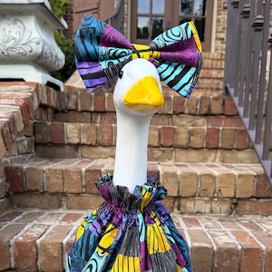May include: A white goose figurine wearing a fabric outfit featuring a patchwork pattern of yellow, purple, blue, and black sections. The goose has a bright yellow beak and wears a matching oversized fabric bow on its head with a ruffled collar.