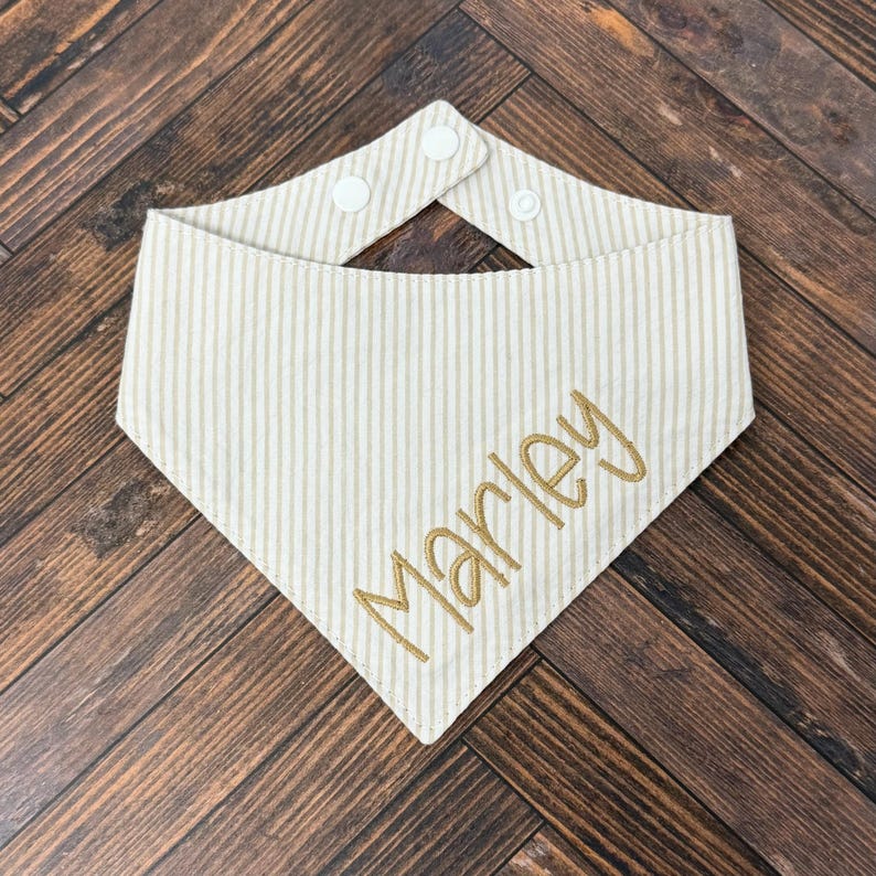 May include: Cream and tan striped pet bandana with the name "Marley" embroidered in gold thread. The bandana has a snap closure at the neck. The background is a dark wood surface.