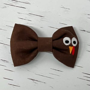 May include: A brown bow tie with a turkey face design. The bow tie features white eyes, an orange beak, and a red wattle. This festive accessory is perfect for Thanksgiving or other special occasions.