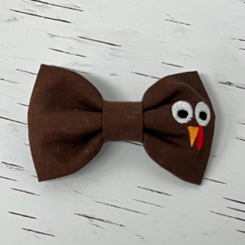 May include: A brown bow tie with a turkey face design. The bow tie features white eyes, an orange beak, and a red wattle. This festive accessory is perfect for Thanksgiving or other special occasions.