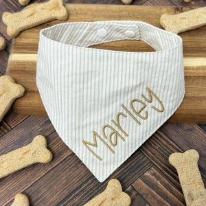 May include: A striped dog bandana in cream and tan with the name "Marley" embroidered in gold thread. The bandana is secured with two white buttons. Dog treats are scattered around the bandana on a wooden surface.