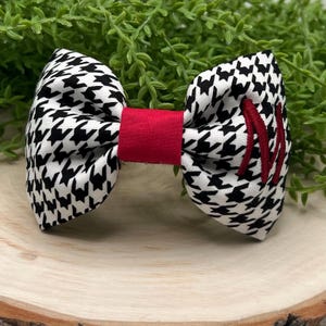 May include: A handmade bow tie featuring a black and white houndstooth pattern. The center band is red, and the right side has red embroidered letters. The bow tie is set against a wooden surface with green foliage in the background.