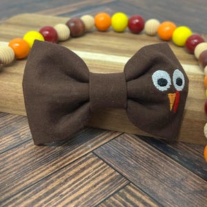 May include: A brown bow tie with a turkey face embroidered on the right side. The bow tie is on a wooden surface with a beaded necklace in the background. The beads are in shades of orange, yellow, red, and brown.